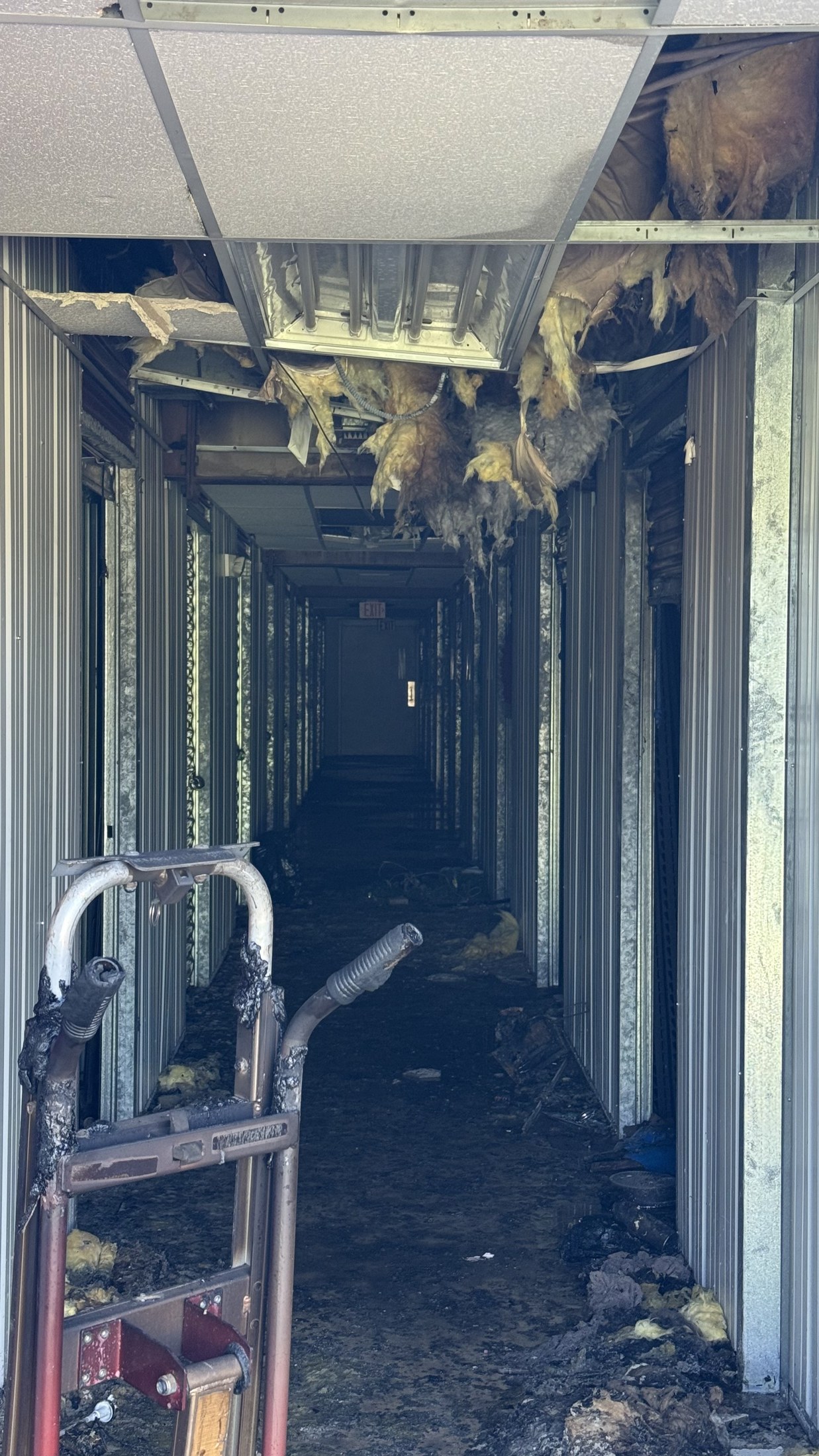 A dark storage-building hallway with damaged ceiling tiles, exposed insulation, and debris on the floor.